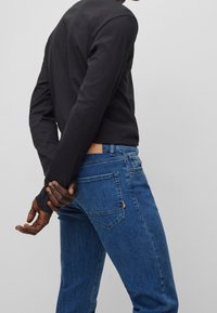 Camisa de manga larga oscura combinada con unos vaqueros de mezclilla azules. Los vaqueros tienen una etiqueta de cuero en la cintura y una sutil textura de desgastado.