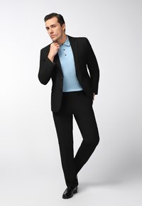 Mann in schwarzanzug und hellblauem Poloshirt, der mit einer Hand am Kinn und der anderen in der Tasche vor einem einfarbigen Hintergrund steht.