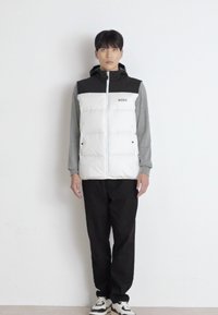 Balta pūkinė liemenė su juoda viršutine dalimi, derinama su pilku ilgomis rankovėmis marškinėliais ir juodomis kelnėmis. Apačioje matomos sportbačiai.