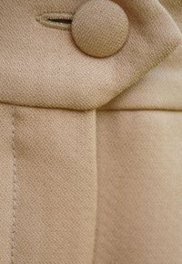 Beżowa tkanina o teksturowanym splocie, z okrągłym zapięciem na guziki i detalami na szwach bocznych, typowymi dla kroju odzieży szytej na miarę.