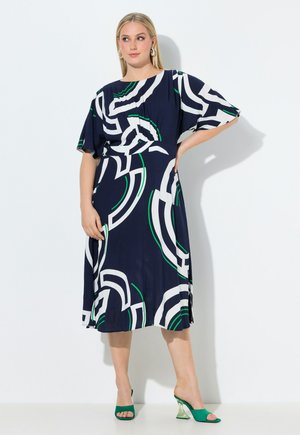 Kvinde i en navyblå midikjole med hvide og grønne abstrakte mønstre, flagrende ærmer, iført grønne peep-toe hæle, stående mod en hvid væg.