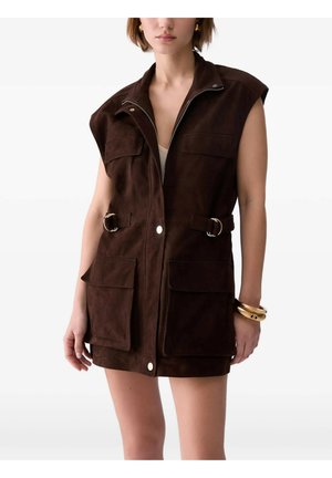 Donna che indossa un vestito gilet senza maniche in camoscio marrone scuro con tasche davanti, cerniera, bottoni a pressione e due cinturini con fibbia in vita