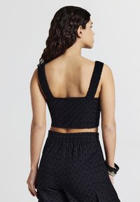 Donna con lunghi capelli ricci che indossa un crop top nero testurizzato con spalline larghe e pantaloni corti a vita alta abbinati, in piedi davanti a uno sfondo semplice.