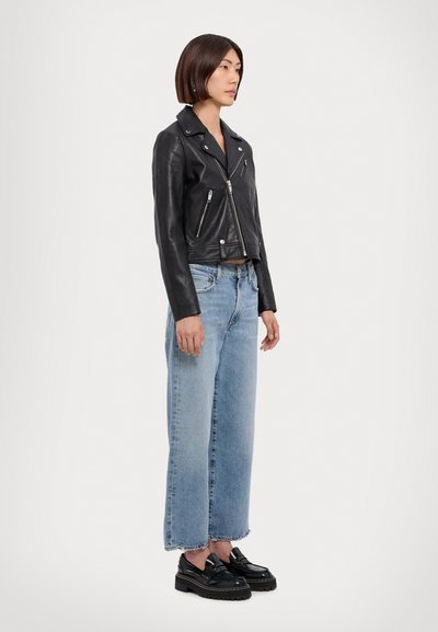 Femme debout, portant une veste en cuir noire, un jean large bleu clair et des mocassins noirs épais, sur fond blanc uni.