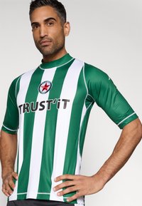 Zielono-biała pasiasta koszulka sportowa z krótkim rękawem, z okrągłym logo z czerwoną gwiazdą i napisem „TRUST IT” na przodzie.
