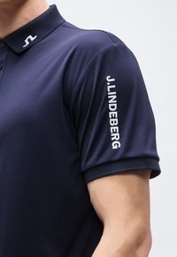 Πουκάμισο πόλο σε ναυτικό μπλε με κεντημένο το J.LINDEBERG στο αριστερό μανίκι και μικρό λογότυπο στο γιακά.