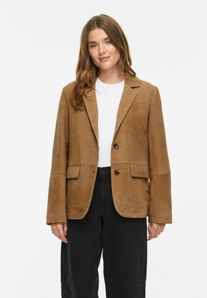 Donna con blazer in camoscio marrone sopra una maglietta bianca e pantaloni neri a vita alta, in piedi su uno sfondo bianco semplice.