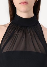 Schwarzes Neckholder-Top aus transparenter Stoff mit hohem Kragen. Verfügt über geraffte Textur und einen unteren Abschnitt aus festem Schwarz. Goldene Ohrstecker sichtbar.