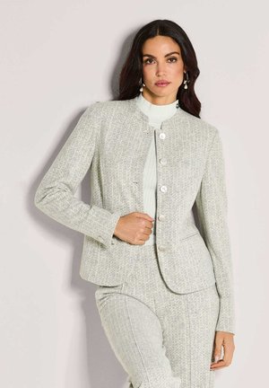 Vrouw met lang, donker haar die een lichtgrijze, gestructureerde, gebutton-down blazer en bijpassende broek draagt, poserend tegen een bleke muur.