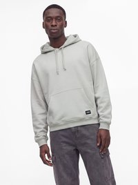 Jeune homme portant un sweat à capuche gris clair avec une poche avant et un pantalon cargo gris foncé, debout sur un fond blanc uni.