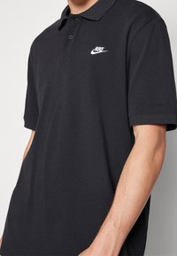 Černé polo tričko z texturované látky, se zapínáním na tři knoflíky a bílým logem Nike na levé straně hrudi. Krátké rukávy.