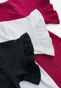 Trīs T-krekli izklāti uz plaknes: viens burgundijas krāsā, viens baltā un viens melnā. Katram ir krokojošu piedurkņu dizains, izgatavots no mīksta kokvilnas auduma.