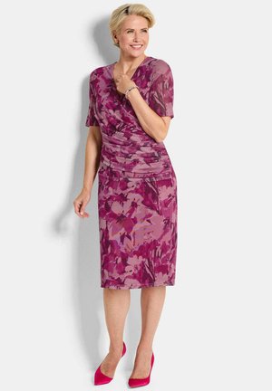 Vrouw die lacht, gekleed in een knielange roze en paarse bloemenjurk met korte mouwen en bijpassende roze hakken, staand tegen een witte achtergrond.