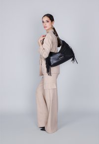 Borsa a spalla in pelle nera con dettagli in frange, abbinata a un outfit in lino beige chiaro composto da pantaloni a gamba larga e una blusa tailored.