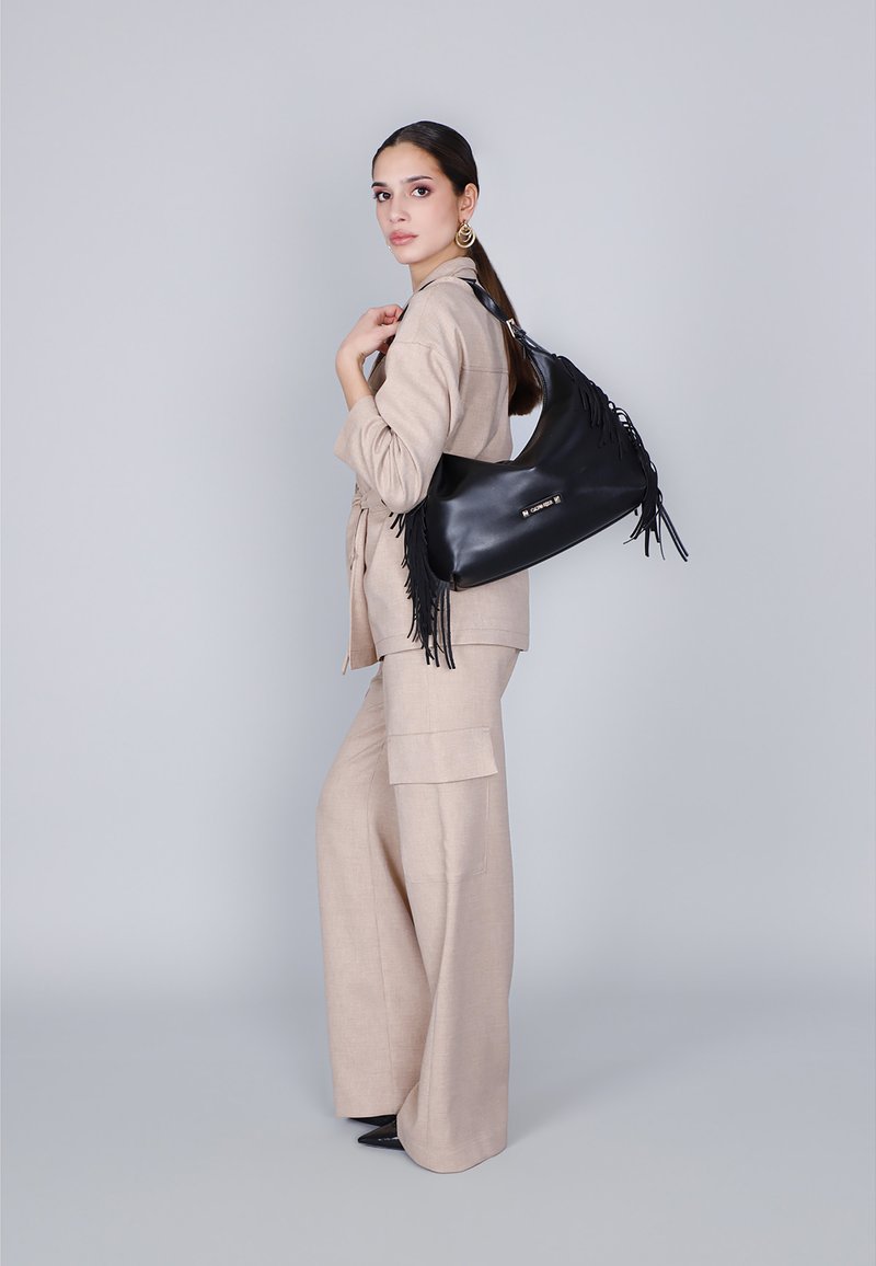 Borsa a spalla in pelle nera con dettagli in frange, abbinata a un outfit in lino beige chiaro composto da pantaloni a gamba larga e una blusa tailored.