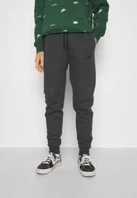 Tummanharmaat hupparit, jotka on valmistettu pehmeästä kankaasta, joissa on slim fit -malli, joustava vyötärö, jossa on nyöri, ja huomaamaton Nike-logo sivulla.