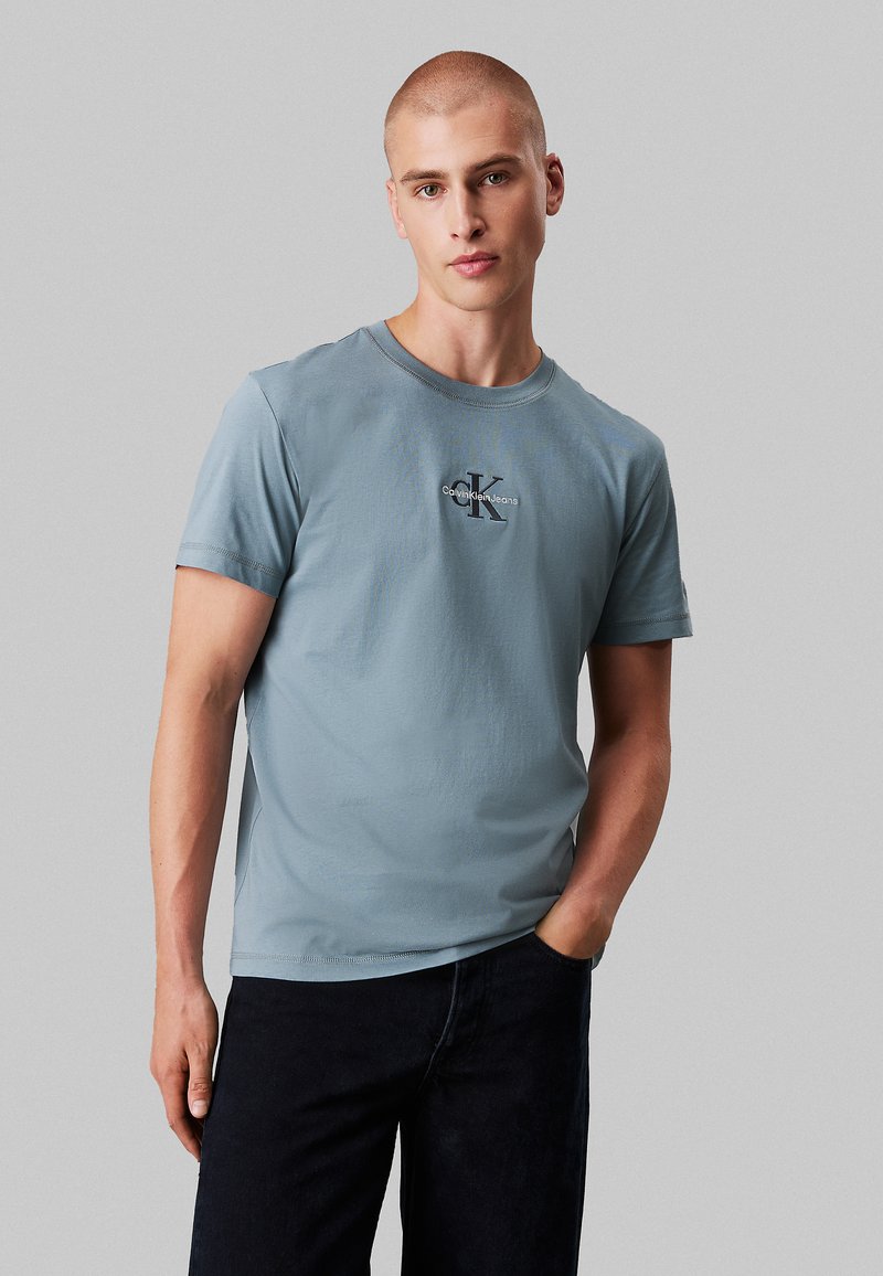 Light blue cotton t-shirt with short sleeves, round neckline, and a central embroidered logo "Calvin Klein Jeans" in dark lettering.