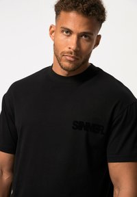 Schwarzes Baumwoll-T-Shirt mit kurzen Ärmeln, versehen mit einem strukturierten, samtigen "SINNER."-Logo auf der linken Brust. Glatte Textur, lockere Passform.