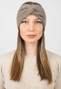 Un bandeau en tricot couleur brun clair, avec un design avant torsadé. Texture douce avec une sensation extensible, complétée par de longs cheveux raides.