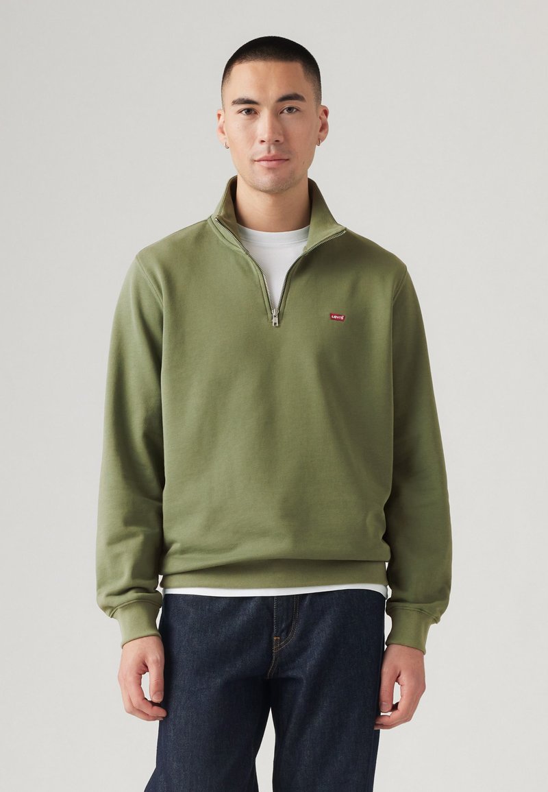 Olijfgroen ritsvest met een hoge kraag, ribbelmanchetten en een klein rood logo op de borst, gecombineerd met donkere spijkerbroeken.