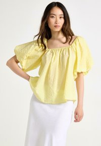 Texturerad gul blus med breda puffärmar och fyrkantig halsringning, stylad med en vit kjol. Tyget har ett subtilt veckat mönster.