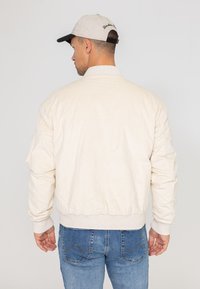 Blouson en velours côtelé beige clair avec des poignets et une taille côtelés. La vue de dos montre une texture lisse et un design minimaliste. Un jean bleu est visible en bas.