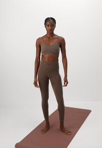 Woman standing barefoot on brown yoga mat, wearing matching brown sports bra and high-waisted leggings against plain background.