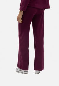 Pantalones holgados color burdeos, confeccionados en una tela suave, con un diseño de pierna acampanada y una textura lisa. Llevados con zapatos blancos y detalles a rayas.