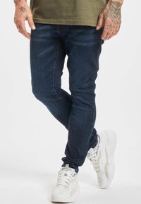 Homme portant un jean slim bleu foncé, des baskets blanches et une chemise vert olive, debout les jambes croisées sur un fond blanc uni.