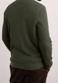 Suéter de punto tipo waffle en verde oliva, con cuello redondeado, dobladillo acanalado y mangas largas, combinado con pantalones de pana oscuros.