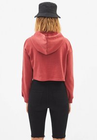 Hoodie rosso accorciato con cappuccio, caratterizzato da maniche lunghe e una texture morbida, abbinato a pantaloni shorts neri in denim a vita alta con orli sfrangiati.