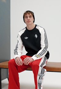 Črno-bela jopa z ogrinjalnimi rokavi z črtami, na kateri je logotip in grafika leva. Rdeče športne hlače z belimi poudarki in logotipi.