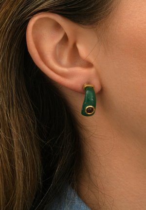 Boucles d'oreilles en résine verte avec un design courbé, présentant un accent en ton doré et une petite pierre précieuse marron incrustée près de la base.