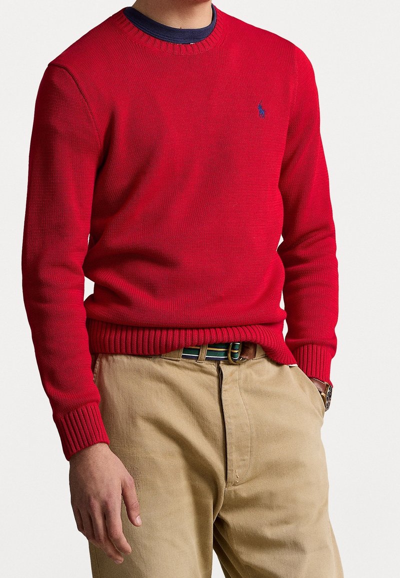 Homme portant un pull en maille rouge par-dessus une chemise bleu marine avec un pantalon beige et une ceinture rayée, main dans la poche sur fond uni.