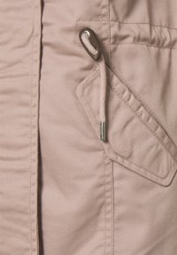 Acercamiento a una tela de color rosa claro que presenta un bolsillo lateral de forma triangular, un ojal metálico y detalle de cordón trenzado.