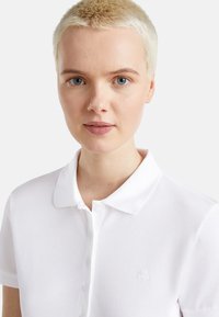 Weißes Polo-Shirt aus strukturiertem Stoff, mit Kragen und kurzen Ärmeln. Mit einem dezenten, gestickten Logo auf der linken Brust.