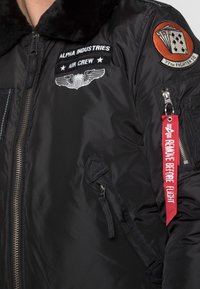 Černá bomber bunda s umělým kožešinovým límcem. Obsahuje nášivky, zipy a červenou značku "Odstranit před letem". Hladký nylonový materiál.