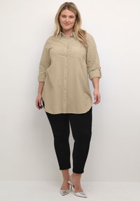 Chemise beige à boutons avec une poche poitrine, manches 3/4 retroussées, associée à un pantalon noir ajusté et des talons argentés. Design simple et classique.