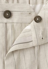 Tissu beige avec de fines rayures verticales, présentant deux boutons marron et un rabat de bouton partiellement ouvert montrant les boutonnières.