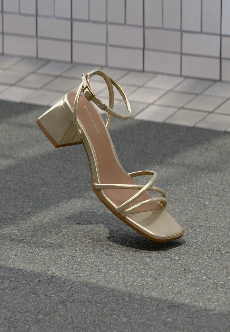Gullfarget sandal med stropper og firkantet hæl, med en glatt, metallisk tekstur og justerbar ankelstropp, plassert på en teksturert overflate.