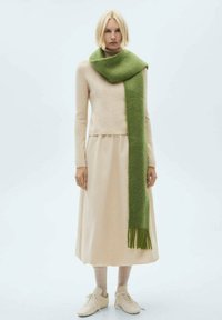Beige sweater, lang grøn tørklæde, beige nederdel; tørklædet har frynser; modellen bærer lyse sneakers og sokker, mens hun står imod en neutral baggrund.