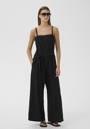 Femme portant une combinaison noire sans manches à jambes larges avec ceinture, lunettes de soleil noires et sandales noires, debout sur fond clair.