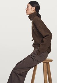 Hnědý žebrovaný rolák s volným střihem, ve spojení s upravenými hnědými kalhotami, sedící na dřevěném stoličce na jednolitém pozadí.