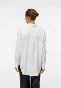 Chemise blanche à manches longues avec col, présentant une coupe droite et des plis dans le dos. Tissu lisse et détails aux poignets.