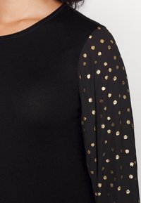 Haut noir avec une texture côtelée et un col rond ; manches transparentes ornées de pois dorés.