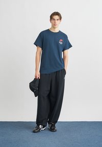 Námořnické tričko s krátkými rukávy, červené logo na hrudi, doplněné volnými černými kalhotami a černými teniskami. V jedné ruce je držena černá čepice.