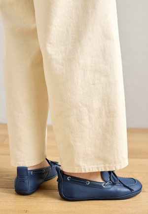 Personne portant un pantalon beige et des mocassins en cuir bleu marine, debout sur un sol en bois à l'intérieur.
