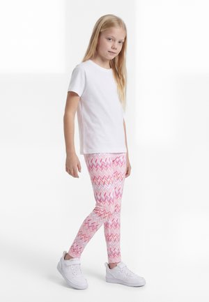 Ung pige iført en hvid kortærmet skjorte, lyserøde og hvide leggings med zigzag-mønster, og hvide sneakers, stående mod en hvid baggrund.