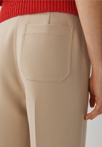 Beige op maat gemaakte broeken hebben een gladde textuur, een vierkante achterzak en zichtbare stikseldetails. Gestoneerd met een rode gebreide top.