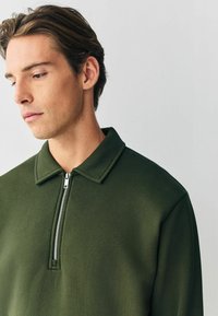 Grüner Reißverschluss-Pulli mit Kragen, der eine glatte Textur und ein minimalistisches Design aufweist. Der Reißverschluss an der Vorderseite reicht bis zum Kragen.